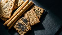 Assorted whole grain and seeded crackers with cinnamon sticks arranged on dark slate surface for food styling, baking content, and culinary backgrounds.