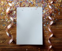 Blank white paper mockup on wooden background with festive pink ribbons and colorful confetti scattered around creating celebratory party atmosphere.