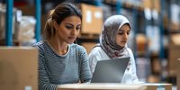Diverse female coworkers collaborate on digital tablet in modern warehouse setting, reviewing inventory data amid storage shelves and cardboard boxes.