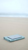 Tablet device resting on folded light blue towel on empty sandy beach with calm ocean and overcast sky, representing digital technology, vacation, relaxation, and remote work.