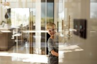Young Caucasian boy with blonde hair peeking from behind glass door in modern home interior with natural lighting and reflections.