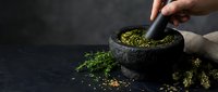 Hand grinding fresh herbs and spices in traditional stone mortar and pestle on dark background for cooking and culinary preparation.