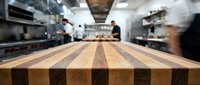Striped wooden butcher block countertop in professional kitchen with blurred chefs working in background, suitable for restaurant and culinary design projects.