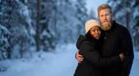 Happy interracial couple embracing in snowy winter forest during golden hour sunset. Romantic outdoor portrait for holiday greeting cards and seasonal campaigns.