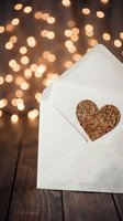 White envelope with gold glitter heart on wooden surface against warm bokeh lights, perfect for Valentine's Day, wedding, love letter, greeting card, and romantic celebration designs.
