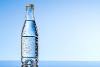 Cold glass bottle with sparkling mineral water and metal cap against blue background, refreshing drink with bubbles and condensation droplets.