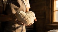 Baker holding fresh artisan bread loaf in warm rustic kitchen with golden sunlight streaming through window creating cozy homemade baking atmosphere.