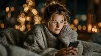 Young blonde woman relaxing at home in cozy sweater using smartphone with warm bokeh lights creating intimate evening atmosphere for lifestyle content.