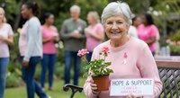 Senior Caucasian woman in pink sweater holding potted flowers at breast cancer awareness event with ribbon symbol and supportive community gathering outdoors.