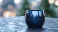 Dark blue ceramic cup with metallic glaze finish on textured surface against blurred bokeh background, creating moody atmospheric composition for beverage and lifestyle content.