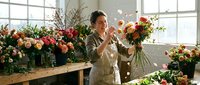 Florist arranging fresh cut flowers in vase at studio workbench with blooming arrangements, natural window light, professional floral design workspace.