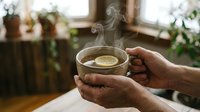 Young adult male with medium-dark skin tone holding steaming cup of herbal tea or hot beverage indoors. Warm, cozy atmosphere with blurred potted plants and window in background. Suitable for wellness, lifestyle, and beverage marketing.
