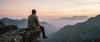 Hiker in camouflage jacket sits on rocky cliff edge overlooking misty mountain valley at sunrise, contemplating vast landscape with birds flying overhead.