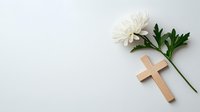 Wooden cross and white chrysanthemum flower on light background. Religious symbol of Christianity with fresh bloom and green leaves for memorial service.
