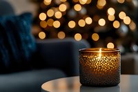 Glowing decorative candle in textured glass holder with warm bokeh lights in background creates cozy atmospheric holiday ambiance for winter season.