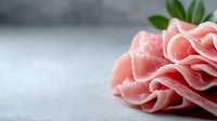 Thinly sliced prosciutto ham arranged in delicate folds on gray surface, macro food photography showing texture and marbling of cured meat. Fresh garnish.
