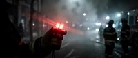 Young male firefighter holding illuminated red emergency light at night scene with colleagues and vehicles in foggy urban street during rescue operation or disaster response.