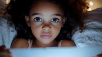 Young African American girl with curly hair illuminated by soft blue light from device screen, creating magical glow while lying in bed at night.