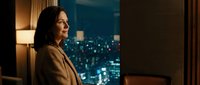 Professional businesswoman in elegant coat standing by window overlooking illuminated city skyline at night, contemplating urban landscape from luxury office.
