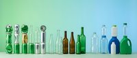 Collection of empty glass bottles in various shapes, sizes and colors arranged in row on white surface against gradient blue-green background for recycling concepts.