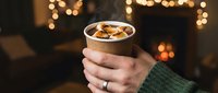 Hands in green sweater holding steaming hot chocolate with toasted marshmallows in paper cup, cozy fireplace and bokeh lights background.
