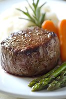 Juicy grilled beef tenderloin steak served with fresh asparagus, baby carrots and rosemary garnish on white plate, close-up view showing meat texture.