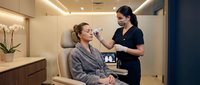 Professional Asian female aesthetician applying eyebrow treatment to Caucasian woman client in modern luxury beauty salon with ambient lighting.