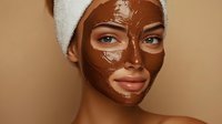 Young mixed race woman wearing chocolate facial mask and white towel on head against beige background, wellness and skincare concept.
