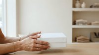 Woman with medium skin tone presenting white stacked boxes on wooden table in bright interior space. Suitable for product packaging, business, retail, and marketing concepts.