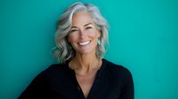 Confident mature woman with silver hair and natural smile wearing black top against turquoise background, representing vitality and professional success in senior years.