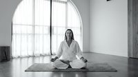 Young woman practicing meditation in lotus position on yoga mat in bright modern studio with large arched window and natural light for wellness and mindfulness.