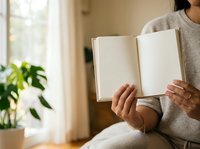 Unrecognizable woman in casual beige sweater holding open blank book near window with green plant, natural daylight, reading and education concept for publishing and learning.