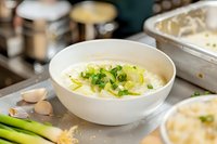 Creamy potato leek soup garnished with fresh chopped green onions and leeks in white ceramic bowl, surrounded by cooking ingredients on rustic kitchen counter.