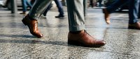 Business professional walking in brown leather shoes on city street with blurred pedestrians in background showing urban commute and corporate lifestyle.