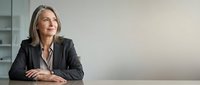 Middle-aged woman with shoulder-length gray hair, Caucasian ethnicity, wearing professional black blazer and white shirt, seated at desk in modern office environment, contemplative expression.