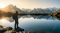 Male hiker with backpack standing by pristine mountain lake at sunrise, snow-capped peaks reflecting in calm water, adventure travel and outdoor recreation concept.