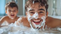 Caucasian father and child having fun bath time moment, playful dad with soap foam beard smiling at camera while toddler laughs in background during family care routine.