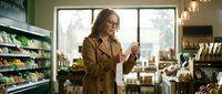 Middle-aged woman with brown hair and glasses wearing a brown jacket, holding a product while shopping in a modern grocery store with shelves and natural lighting.