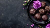 Dark chocolate truffles with purple flowers on black plate against textured background, gourmet confectionery with elegant presentation for dessert menu photography.