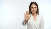 Professional businesswoman in white suit making stop gesture with raised hand against neutral background for corporate communication concepts.
