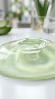 Clear gel serum or cosmetic essence with water droplets on white surface, surrounded by skincare products and green plants in blurred background for beauty and wellness.