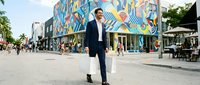Businessman in dark suit walking through urban street with colorful mural wall art, modern city district with pedestrians and cafes, professional man commuting in contemporary downtown area.