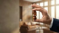 Businessman holding wooden house miniature in modern apartment interior with natural light from windows.