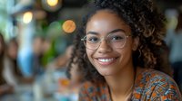 Young African American woman with curly hair and glasses smiling warmly at camera. Natural bokeh background creates soft, professional atmosphere in office or cafe setting.