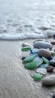 Colorful sea glass and smooth pebbles scattered on sandy beach with gentle ocean waves washing ashore creating peaceful coastal scene for relaxation concepts.