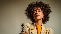 Young woman with curly dark hair and medium brown skin tone wearing a mustard yellow top and beige blazer, smiling confidently at camera for professional portrait or business use.