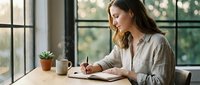 Young professional woman writing in notebook at wooden desk by large windows with natural light, focused on work planning and productivity.