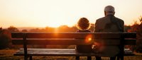 Senior couple sitting on park bench watching golden sunset together, romantic evening moment, retirement lifestyle, peaceful outdoor scene, love and companionship.