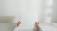 Two hands in white long-sleeved shirt holding blank white paper sheet on wooden table with neutral background for mockup and branding purposes.