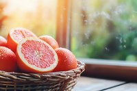 Fresh pink grapefruit halves and whole fruits in wicker basket near window with natural sunlight and water drops, morning breakfast setting.